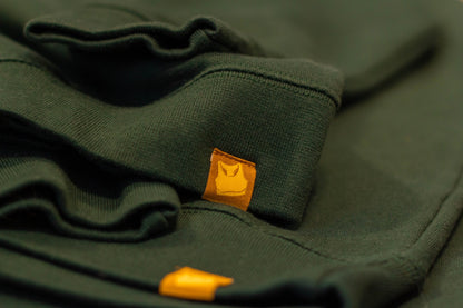 The Sports Bra pinch label tag close-up. Sweatshirt looks soft, is evergreen color, and the pinch label is a burnt ochre color with the Sports Bra official symbol on the tag. There is a nice contrast. 